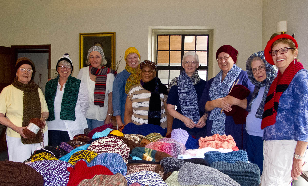 Nine Serendipity Stitchers are smiling with their hats and scarves at a meetup.